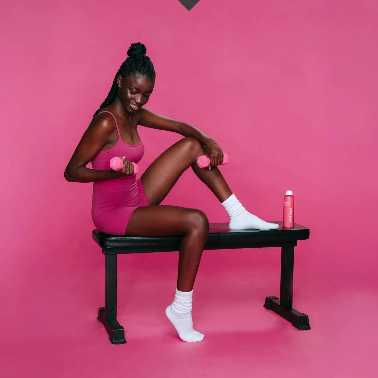 Eine Frau in einem rosa Outfit sitzt auf einer Bank und hält zwei rosa Hanteln. Neben ihr liegen Clarifying Body Spray + Daily Microfoliant, alles vor einem leuchtend rosa Hintergrund.