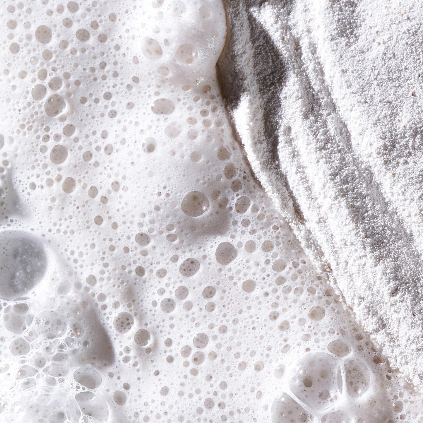 Nahaufnahme von Mehl und schaumiger Flüssigkeit – Blasen auf der einen Seite und pudrige Textur auf der anderen Seite erinnern an Clarifying Body Spray + Daily Microfoliant, bereit, Ihre Haut zu erfrischen, zu peelen und zu klären.