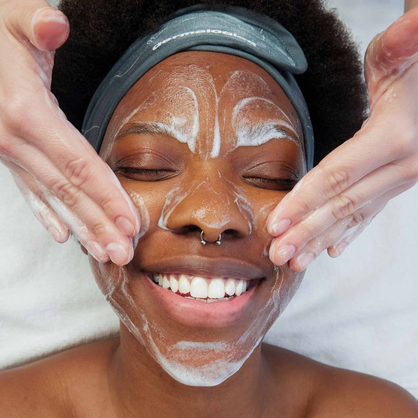 Die Person lächelt mit geschlossenen Augen, während sie eine Gesichtsbehandlung erhält; die Hände tragen Daily Microfoliant + Special Cleansing Gel auf und erzeugen einen schaumigen Schaum auf dem Gesicht.