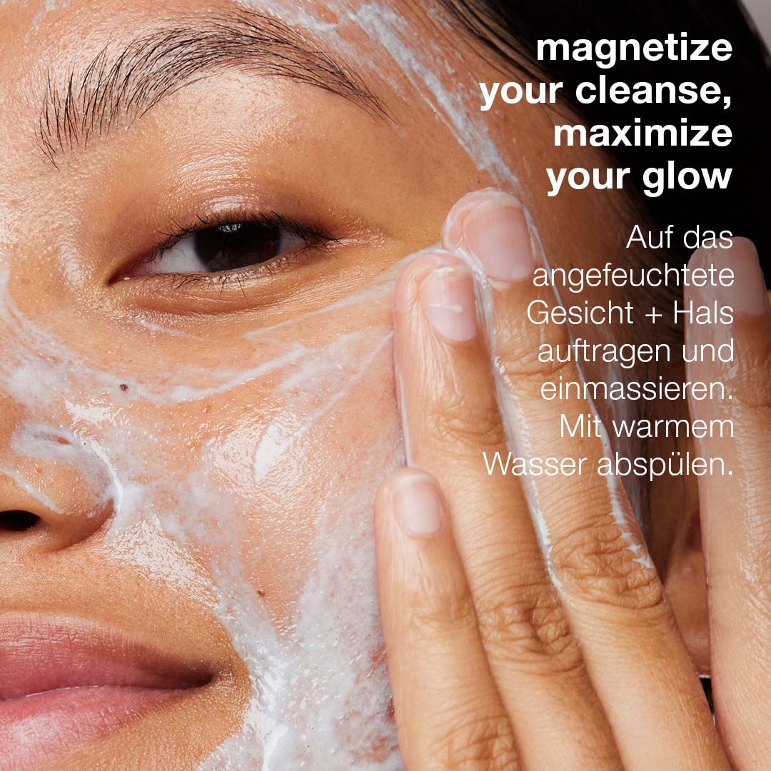 Eine Person verwendet Magnetic[+] Afterglow Cleanser – Review und massiert die cremige, feuchtigkeitsspendende Formel in ihr Gesicht ein; Anweisungen werden sowohl auf Englisch als auch auf Deutsch bereitgestellt.