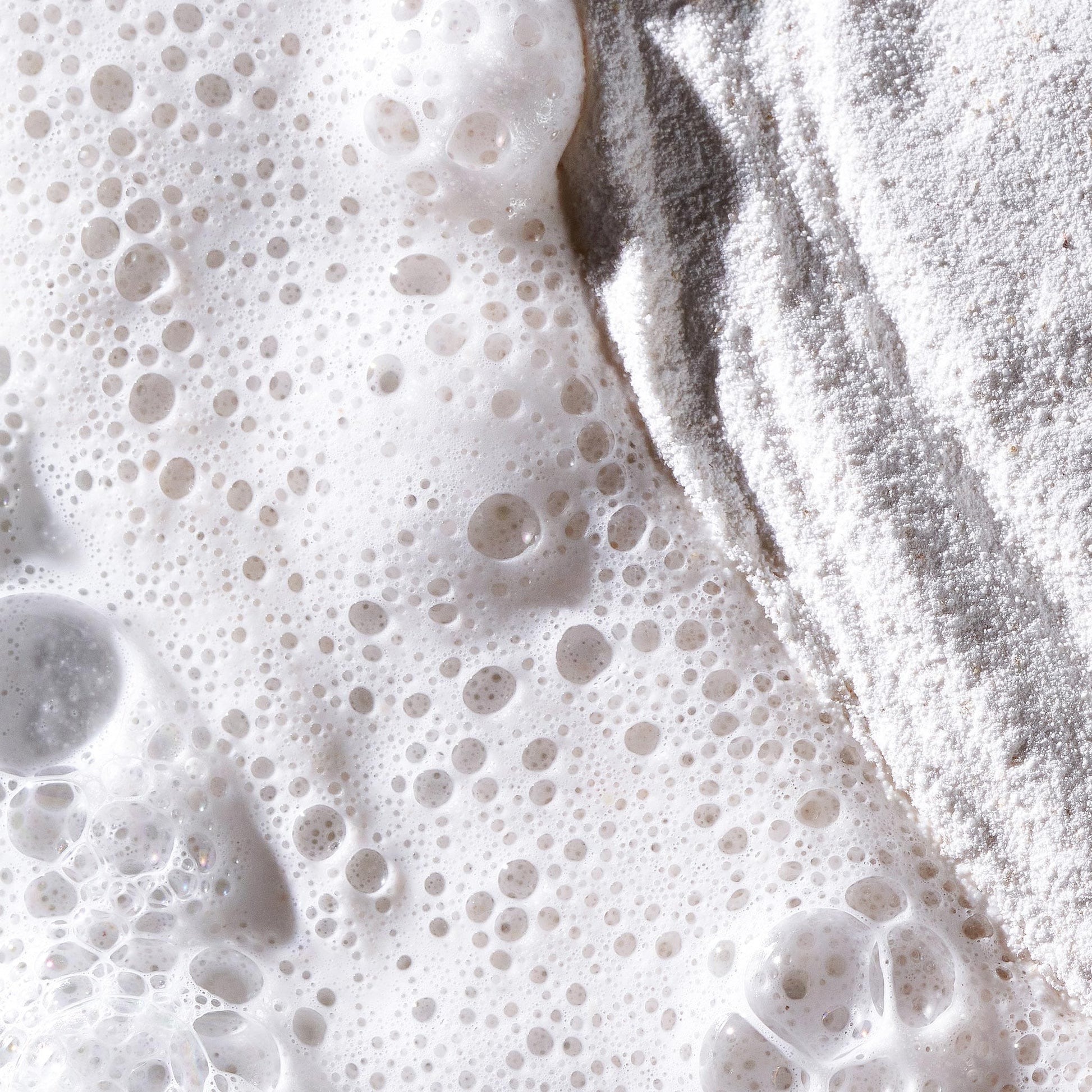 Nahaufnahme von Mehl und schaumiger Flüssigkeit – Blasen auf der einen Seite und pudrige Textur auf der anderen Seite erinnern an Clarifying Body Spray + Daily Microfoliant, bereit, Ihre Haut zu erfrischen, zu peelen und zu klären.