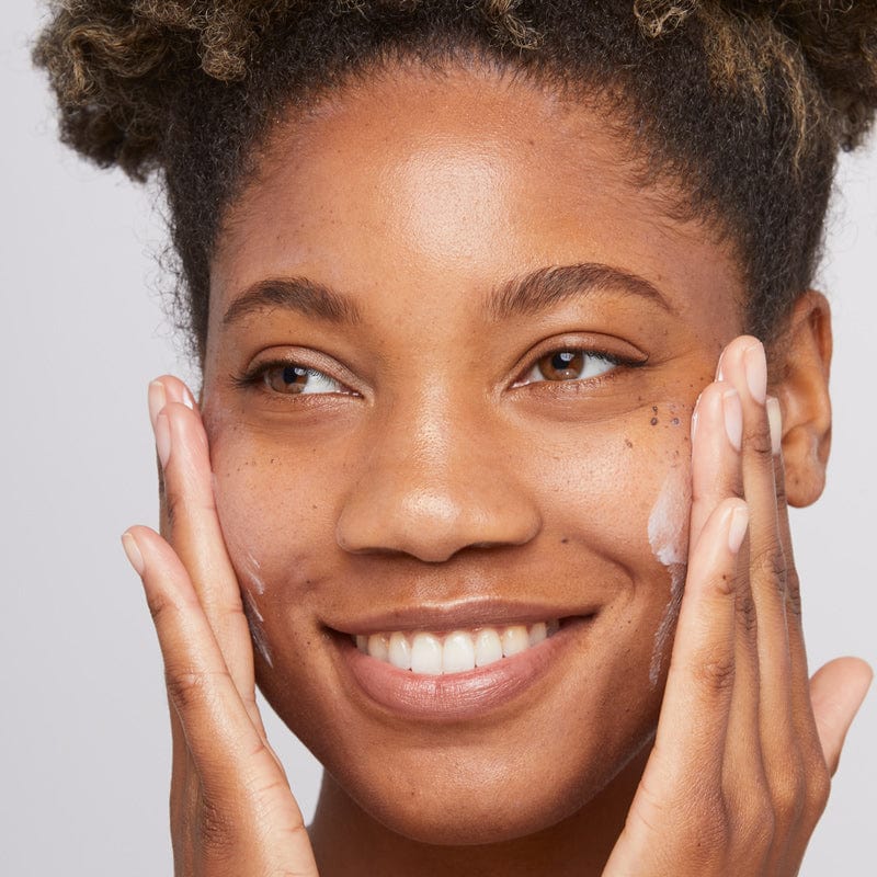 Eine Person lächelt, während sie die Skin Smoothing Cream + Daily Microfoliant mit beiden Händen auf ihre Wangen aufträgt.