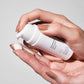Hände drücken eine Lotion oder ein Serum aus einer weißen Flasche in die Handfläche.